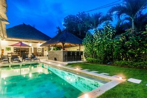 Un área de piscina con bar y sillones de descanso, rodeada de vegetación exuberante y una estructura con techo de paja.