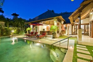 A pool area with lounge chairs and umbrellas, a thatched-roof building, and a well-lit pathway. - Villa Olive (Seminyak)