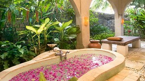 A spa area with a large oval bathtub filled with water and pink flower petals, surrounded by lush tropical plants and a stone bench with a cushion.