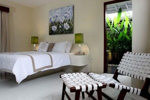 A hotel room with a bed, two bedside tables, a chair, a painting on the wall, and a view of greenery through a glass door.