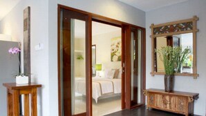 A hotel room with a sliding glass door leading to a bedroom, a wooden console table, and a mirror with a decorative frame. - Villa Padar (Kerobokan)