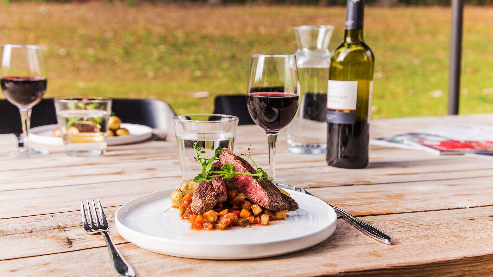 Een bord steak met groenten op een houten tafel, omringd door wijnglazen en flessen.