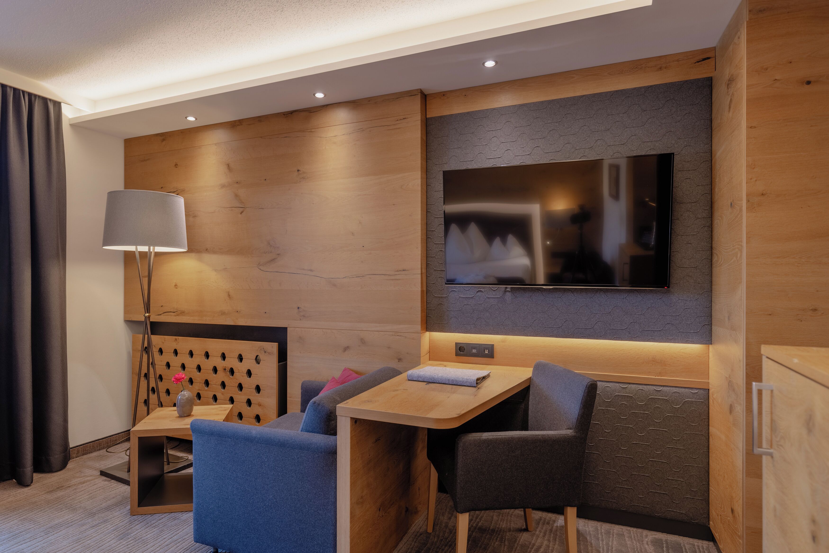 Ein modernes Hotelzimmer mit einer Wandpaneele aus Holz, einem flachbildfernseher über einem Holztisch, einem Sofa und einem Schreibtisch mit Lampe.
