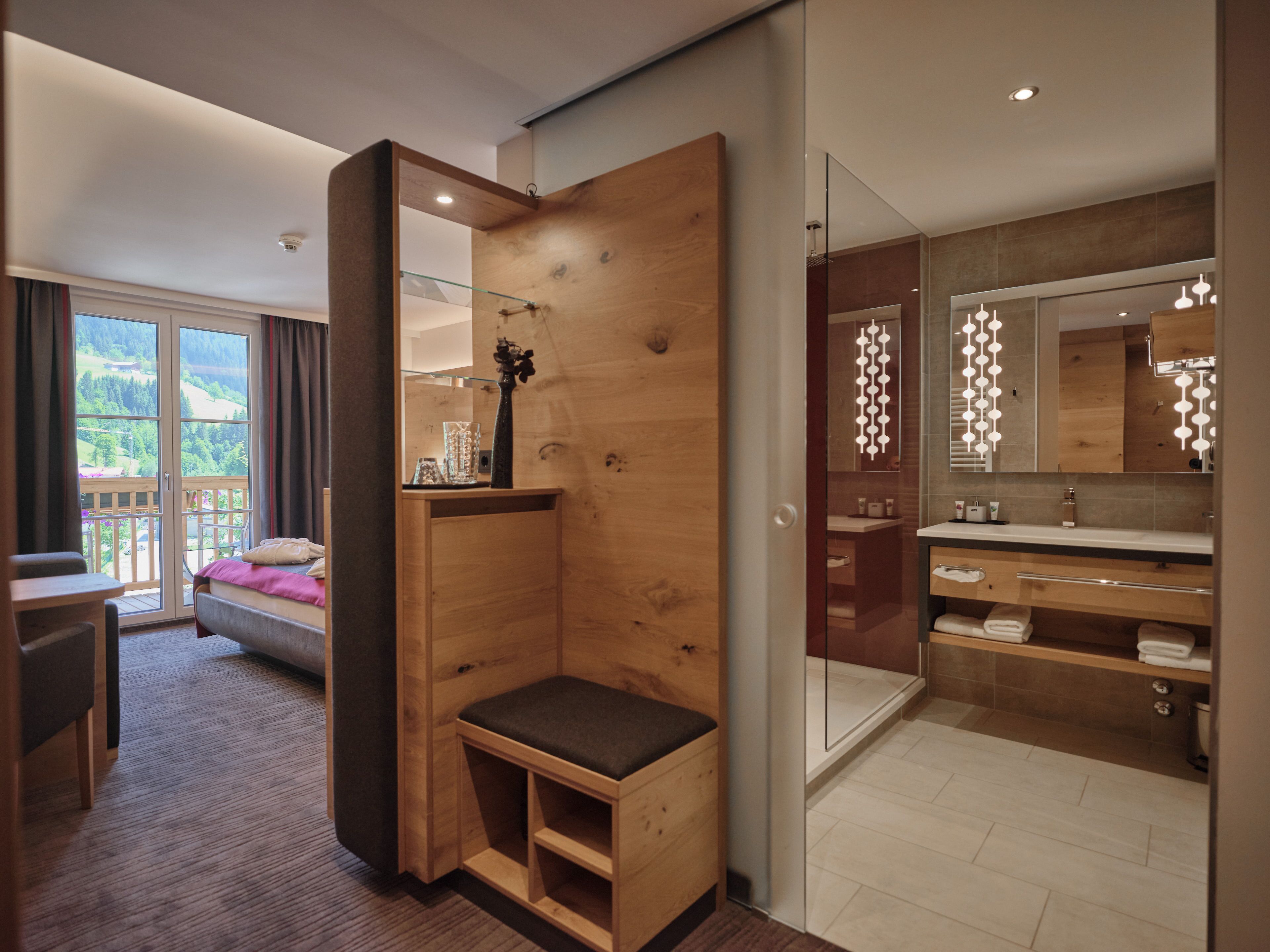 Quarto de hotel moderno com um banco de madeira, um espelho grande, um banheiro com toucador e uma varanda com vista para as montanhas.