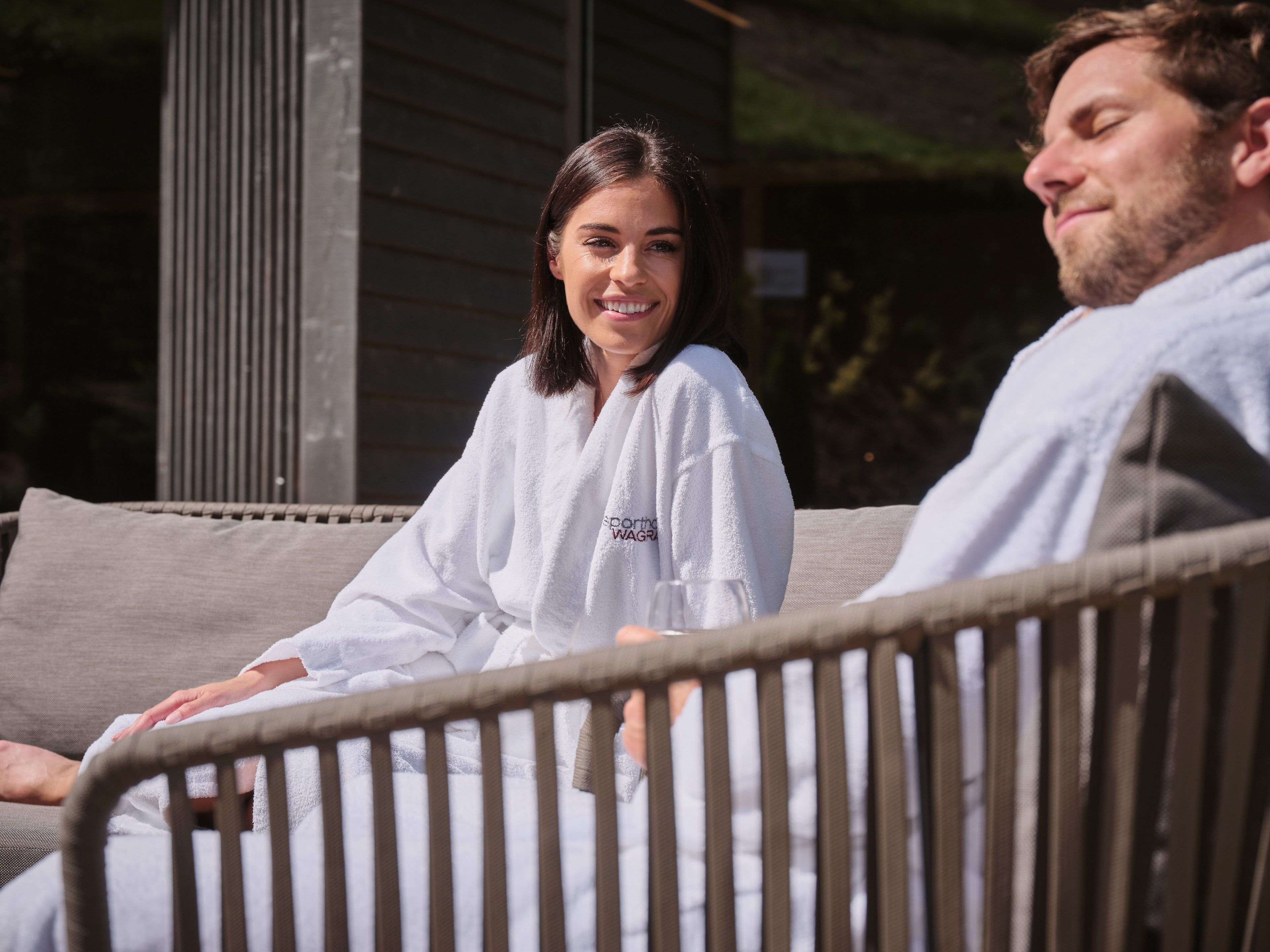 A couple sitting on a wicker sofa outdoors, with a wooden structure in the background.