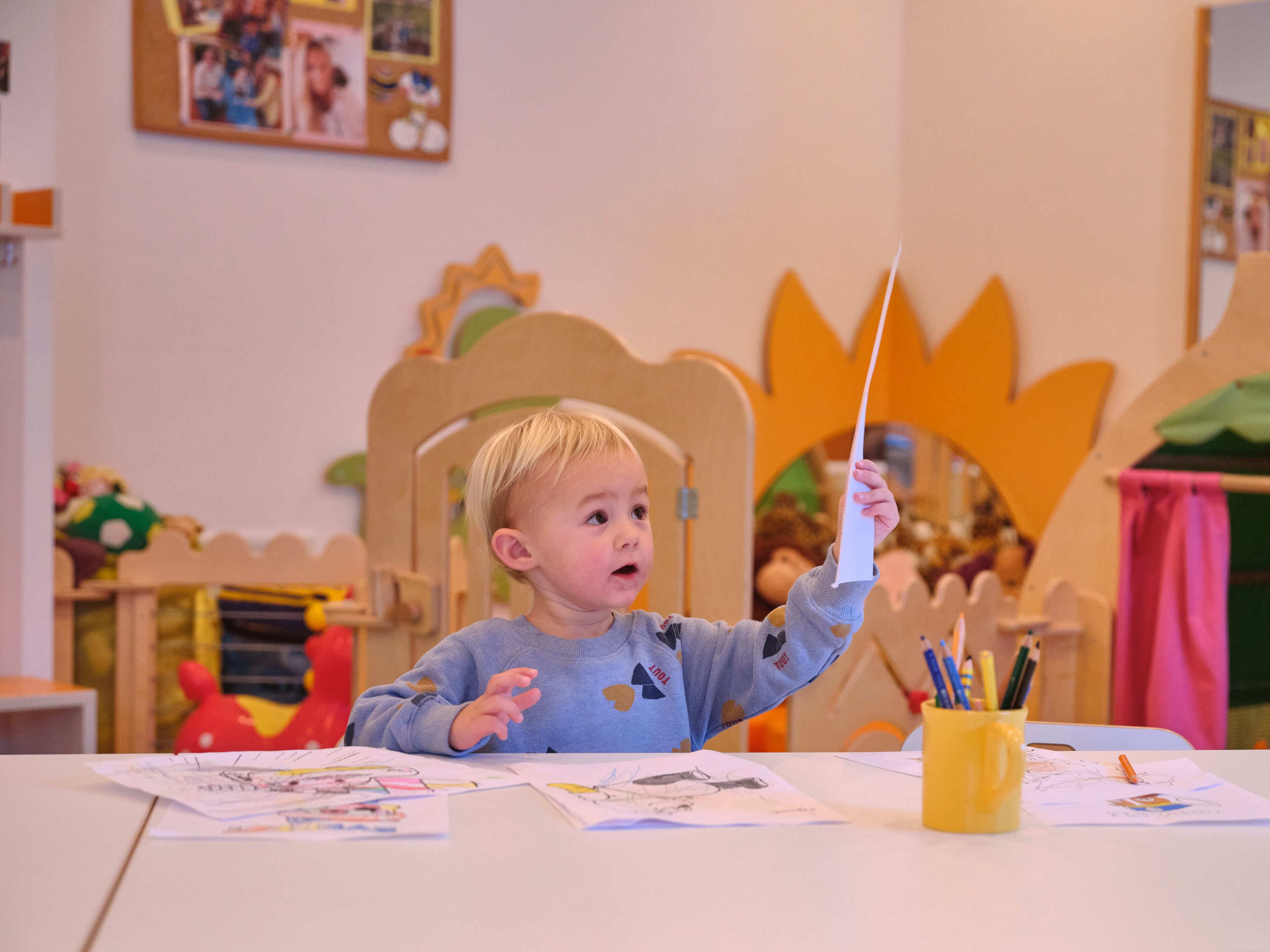 Een kind aan een tafel met papieren en een gele mok, in een kamer met een houten stoel en een grote oranje decoratie.