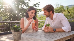 Two people sitting at an outdoor table with drinks, surrounded by greenery and a railing. - Sporthotel Wagrain (Wagrain)