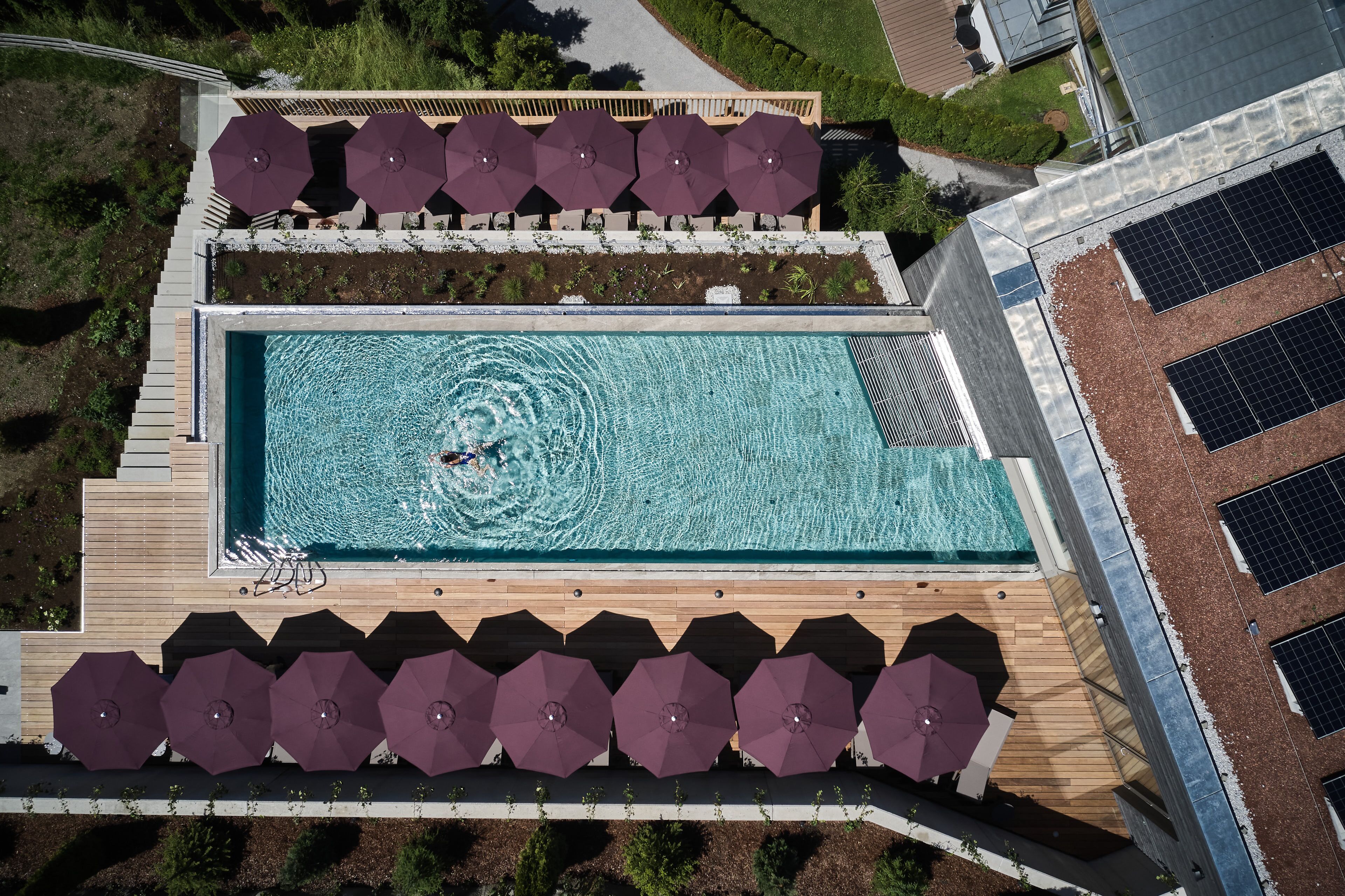 An aerial view of a rooftop swimming pool with surrounding sun loungers and umbrellas, and adjacent solar panels.