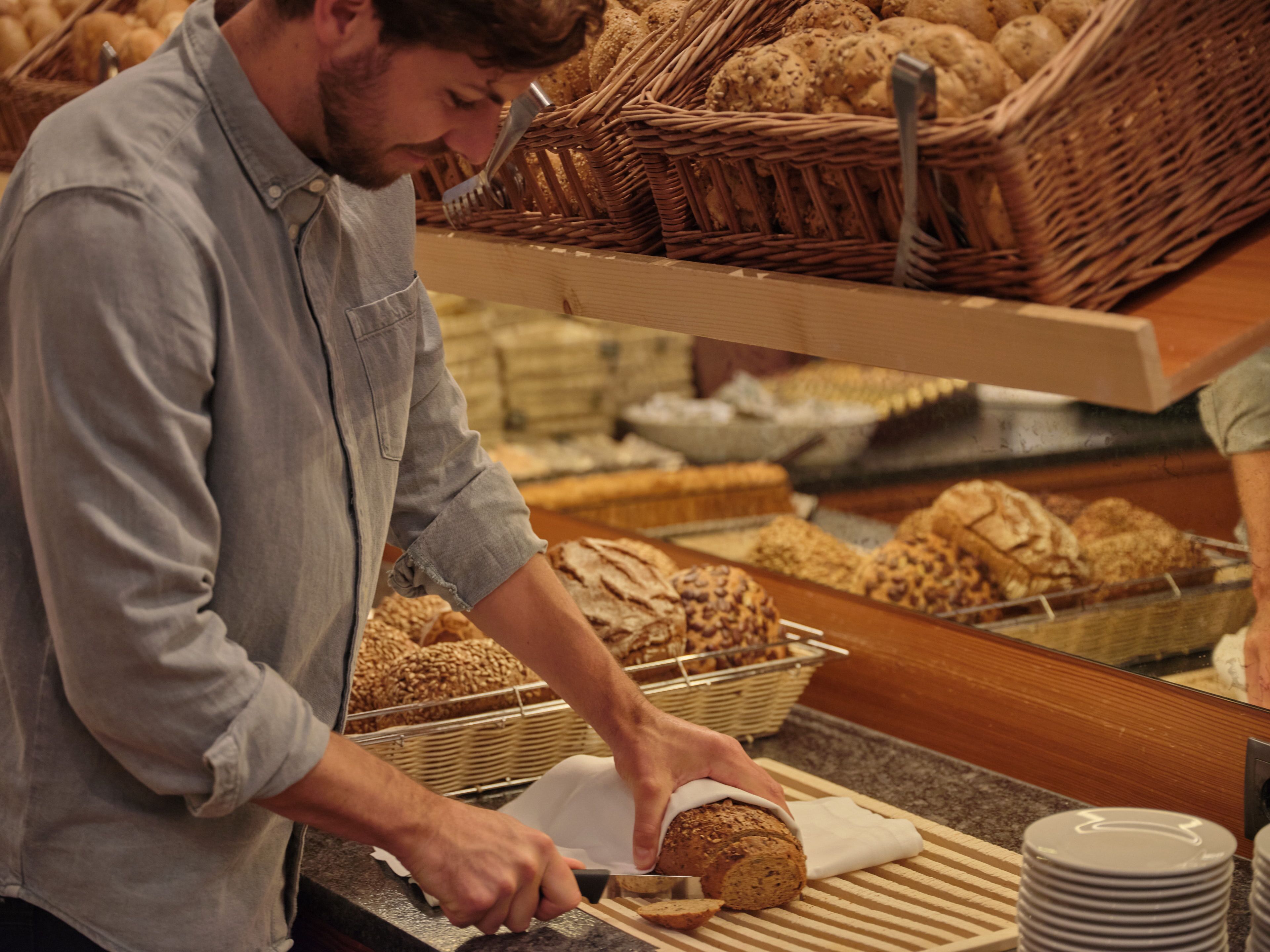 Une personne est en train de trancher du pain sur un comptoir, avec différentes sortes de pain et des paniers en arrière-plan.