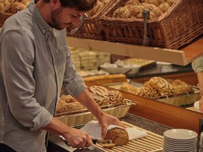 A person slicing bread on a counter with various breads and baskets in the background. - Sporthotel Wagrain (Wagrain)
