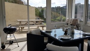 Een eetgedeelte met een glazen tafel, zwarte rieten stoelen en uitzicht op een tuin en gebouwen door grote ramen.