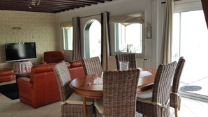 Een woonkamer met een leren bank, rieten stoelen, een houten eettafel en een flatscreen televisie tegen een bakstenen muur.