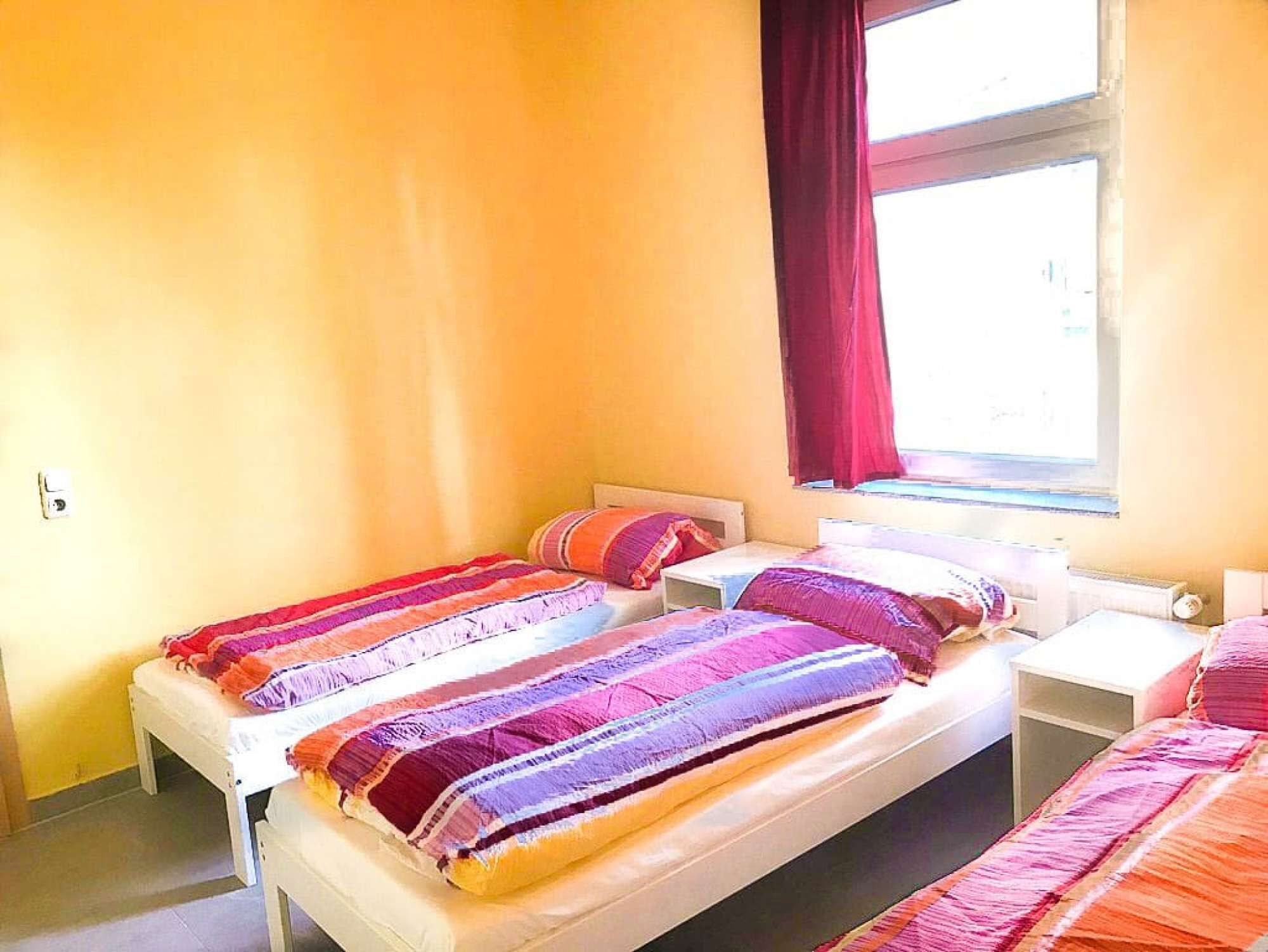 A room with two beds, each with colorful bedding, a window with red curtains, and a white wall.