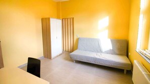 A small, bright room with yellow walls, a single bed, a wooden wardrobe, and a radiator. - T1-Apartment-1.OG-HH (Bergisch Gladbach)