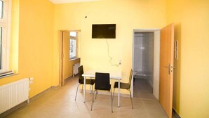 A room with yellow walls, a white table with two chairs, a TV mounted on the wall, and a door leading to another room. - T1-Apartment-1.OG-HH (Bergisch Gladbach)