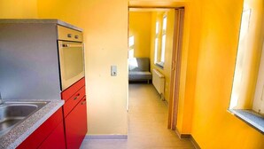 A narrow hallway with a kitchen area on the left, featuring a red cabinet and a stainless steel sink. The walls are painted yellow, and there is a window with a curtain on the right side. - T1-Apartment-1.OG-HH (Bergisch Gladbach)