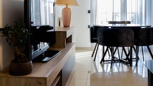A modern living room with a flat-screen TV, wooden shelves, a lamp, and a dining area with a table and chairs. - AR apartment (Πλατανίδια)