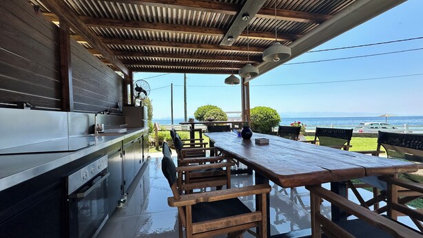 An outdoor dining area with a wooden table and chairs, a kitchen area with a stainless steel countertop, and a view of the sea. - AR apartment (Πλατανίδια)