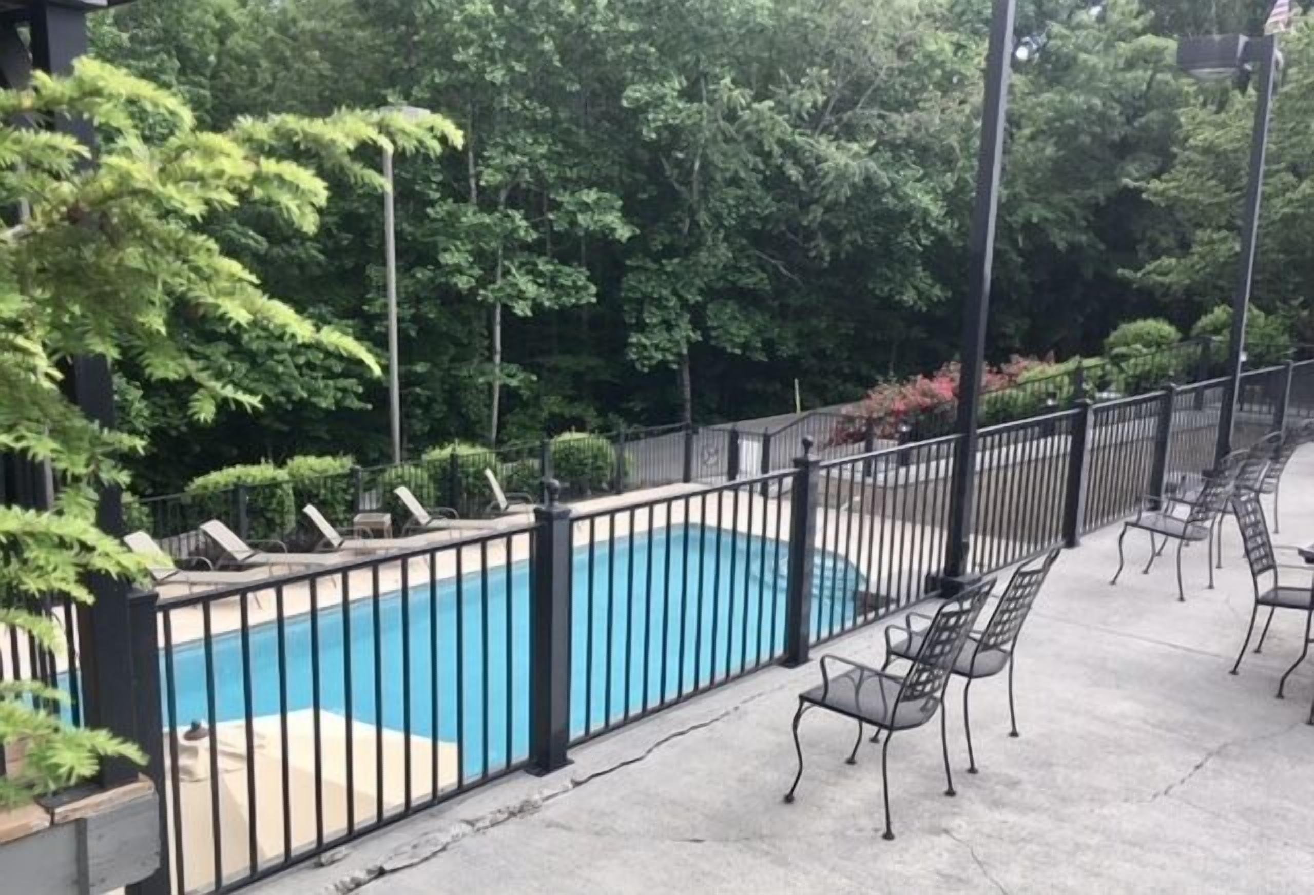 A swimming pool with a black metal fence, surrounded by trees and a concrete deck with metal chairs.