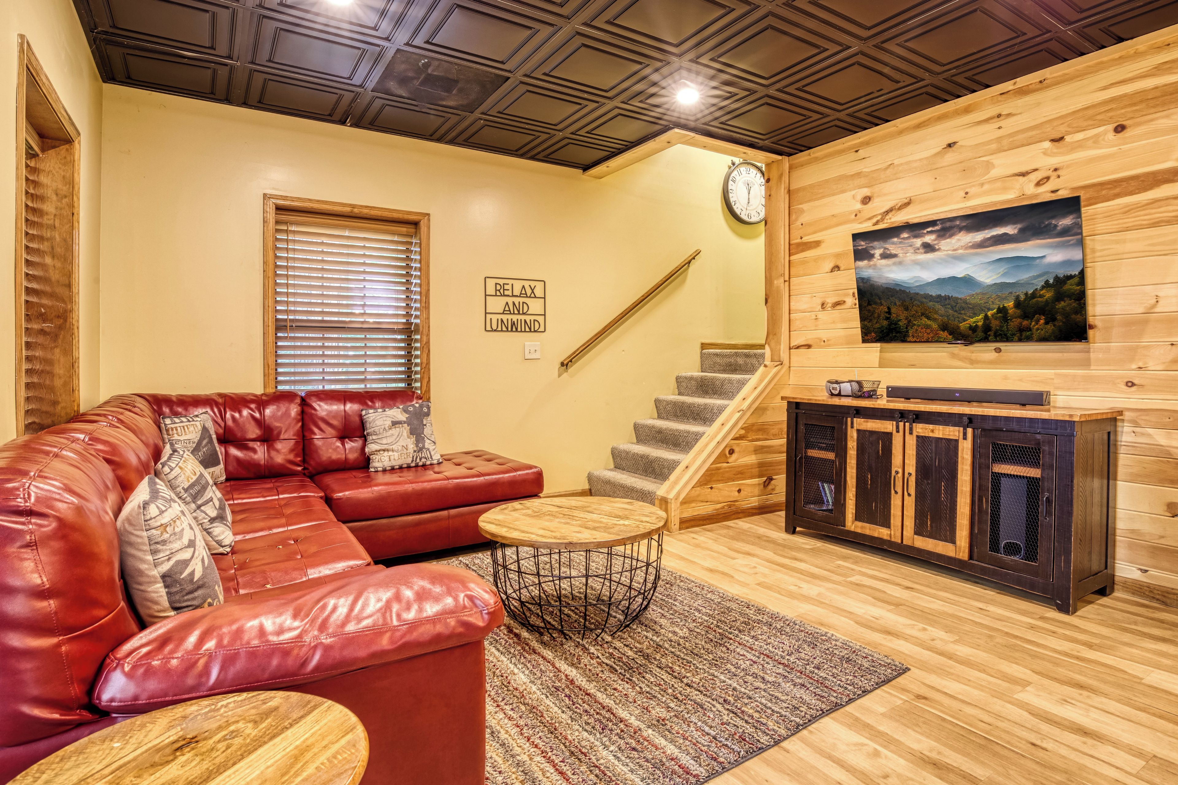 A cozy living room with a leather sofa, a wooden staircase, a TV mounted on the wall, and a scenic landscape painting.
