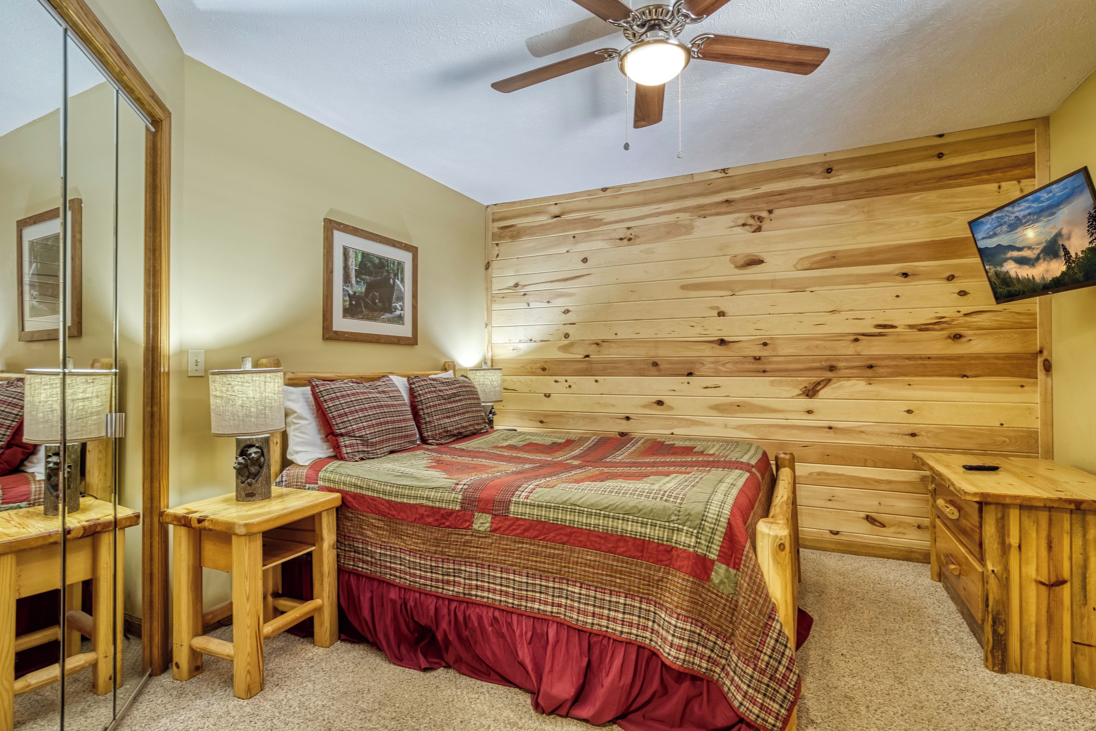 Un dormitorio con cabecera de madera, cama con colcha verde y roja, mesita de noche con lámpara, espejo y un cuadro en la pared.