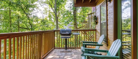 Una terraza de madera con una parrilla, dos sillas Adirondack y vistas a un bosque.