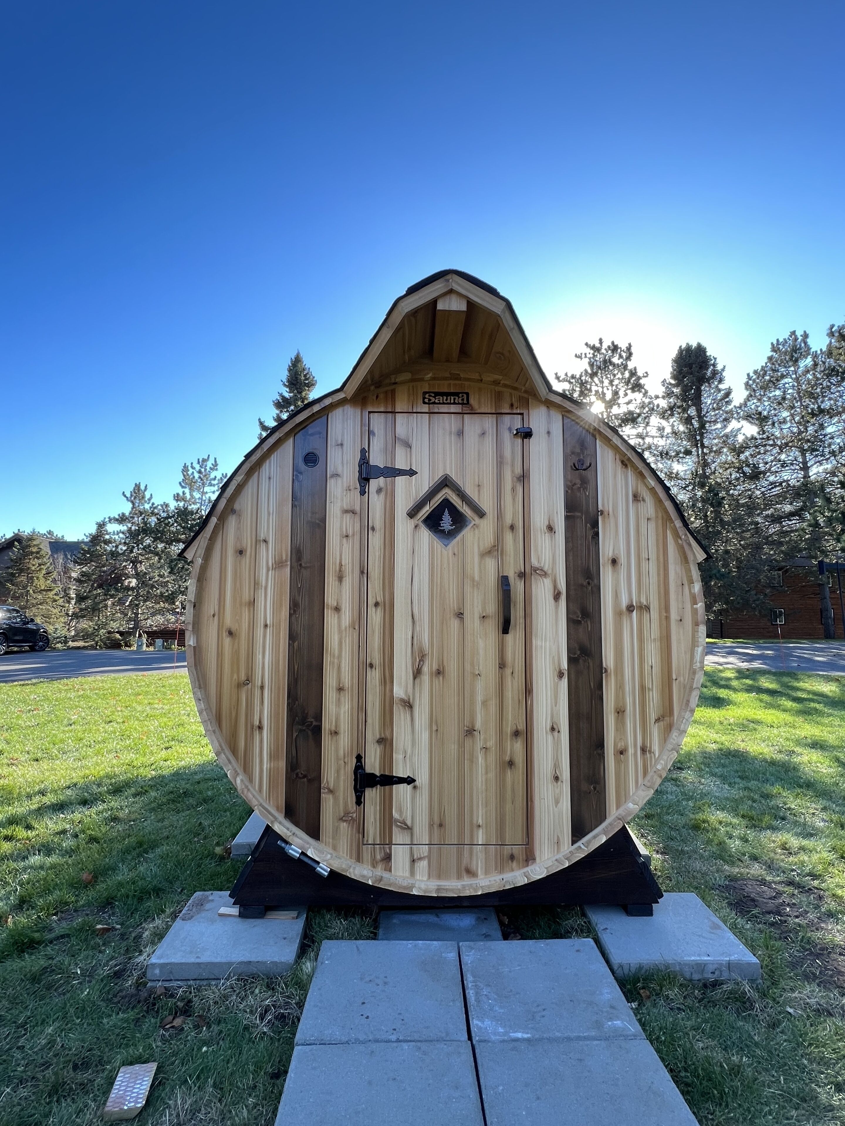 Eine Holzsauna mit spitzer Dachform, einem diamantförmigen Wappen und einem Schild mit der Aufschrift 'Sauna' auf einer Rasenfläche mit einem gepflasterten Weg.