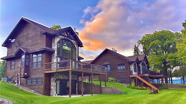 A large, multi-story wooden house with a stone facade, a balcony, and a well-maintained lawn. - Bay Lake Cabin w/200ft of Lakefront, fun deck, private dock + woodfire sauna!  (Deerwood)