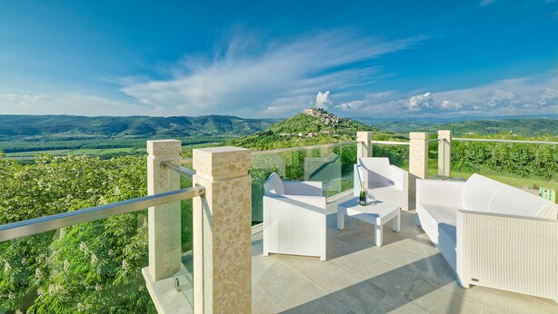 Une terrasse dotée de meubles en osier blanc, d’une balustrade en verre et offrant une vue panoramique sur une colline surmontée d’un bâtiment.