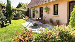 Une maison avec un patio, une table à manger et des chaises. On y trouve également des plantes en pot et une pelouse.