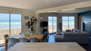 Un salon moderne doté d’une grande fenêtre donnant sur la plage, un coin repas avec une table en bois et un espace salon confortable avec un canapé.