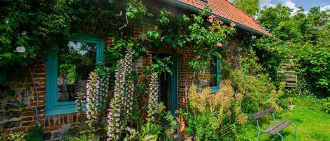 Una casa de ladrillo rústica con tejado de tejas rojas, contraventanas verdes y un jardín con diversas plantas y un banco.