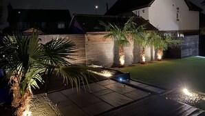 A residential property at night with a well-lit patio area, a swimming pool, and a white house with a dark roof.