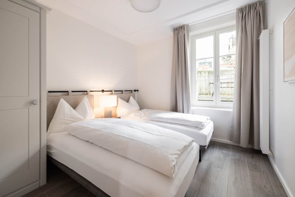 Ein Schlafzimmer mit zwei Betten, einem Fenster mit Vorhängen, einem Heizkörper und einer Tür.