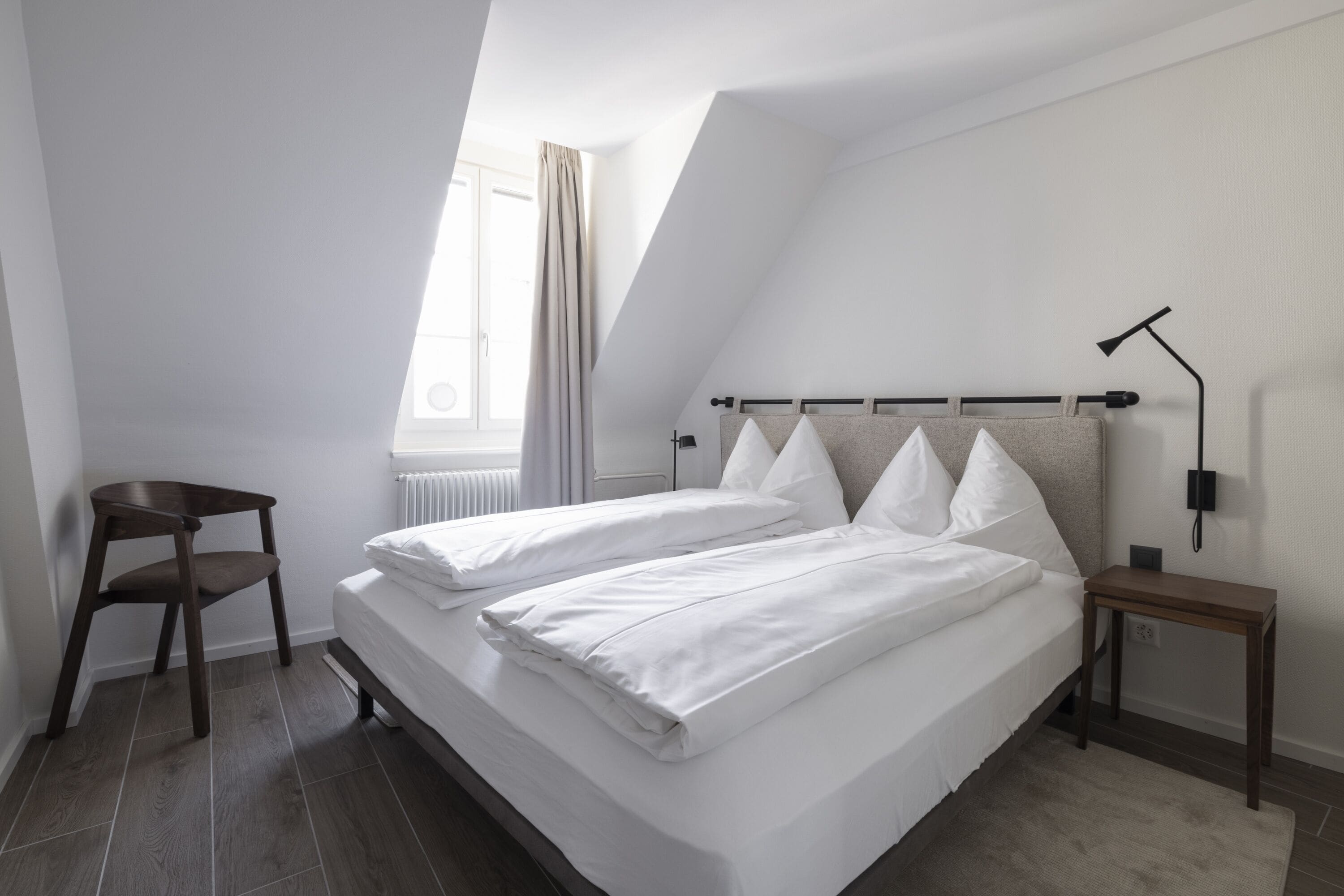 A modern bedroom with a loft-style bed, a small wooden chair, a bedside table, and a window with curtains.