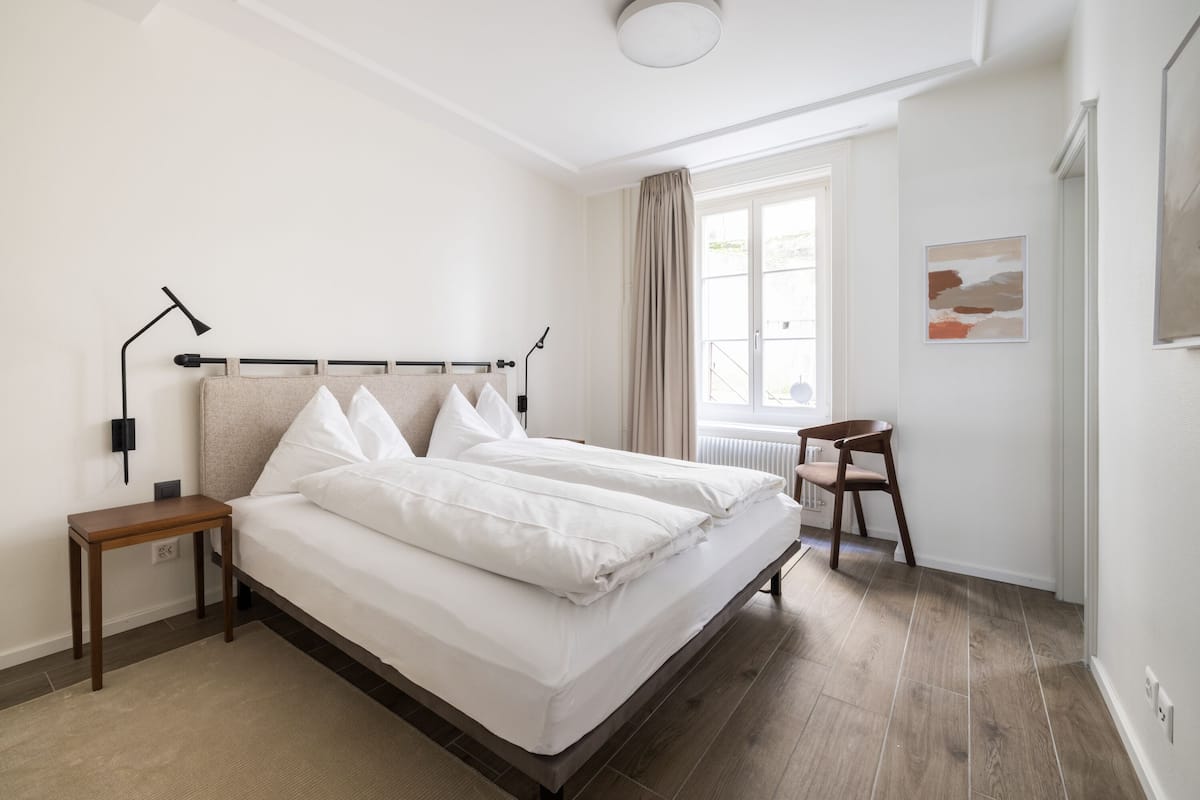 Ein Schlafzimmer mit Bett, Nachttischen, einem Sessel, einem Fenster mit Vorhängen und einer Heizung.