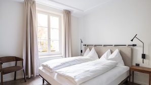 Un dormitorio con cama, una silla, una mesita de noche, una lámpara y una ventana con cortinas.