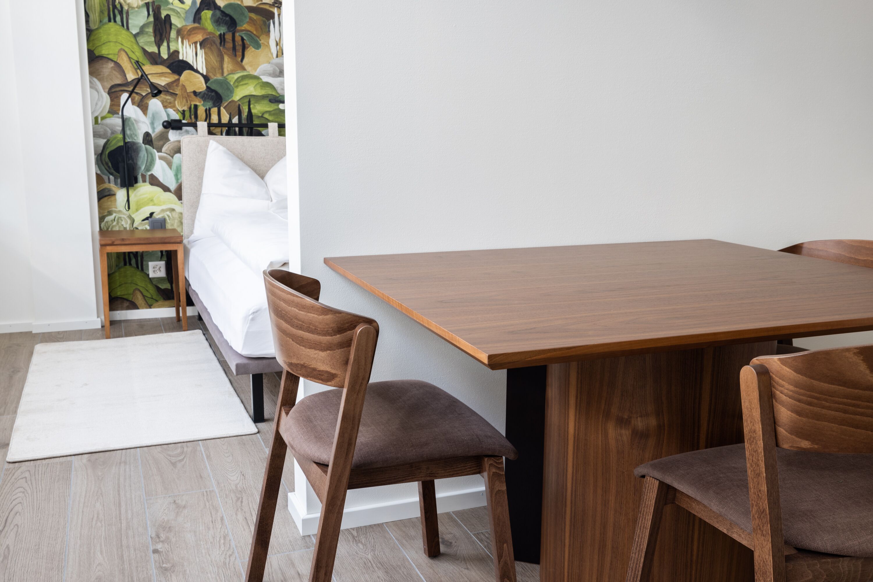 Ein moderner Essbereich mit einem Holztisch und Stühlen, einem weißen Teppich und einem Bett, dessen Wand hinter dem Bett einen grün-weißen Musteraufdruck hat.
