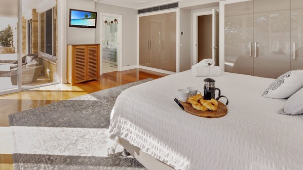 A modern bedroom with a large bed, a TV, a wardrobe, and a bathroom visible through a glass door.