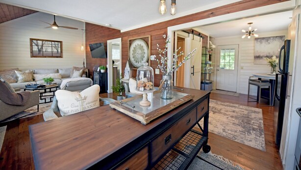 A spacious living room with a wooden dining table, a sofa, and a large mirror. There are decorative items on the table and a ceiling fan above. - Creekside Cottages : Sneffles Mountain Luxury in Town (Blairsville)