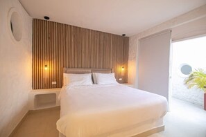 A modern bedroom with a large bed, wooden panel wall, two bedside lamps, and a view of a beach through an open door.