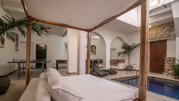 Una cama con dosel con ropa de cama blanca en una habitación con piscina, sillones de descanso y zona de comedor.