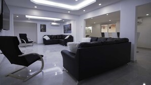 Una sala moderna con un sofá modular negro, un sillón negro y un sofá blanco. Hay dos cuadros en la pared, un televisor de pantalla plana montado y un techo con iluminación empotrada.