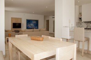 A modern living room with a large flat-screen TV, a painting, a sofa, and a dining area with a wooden table and chairs.