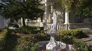 A garden with a fountain, a white fence, and a building with columns.