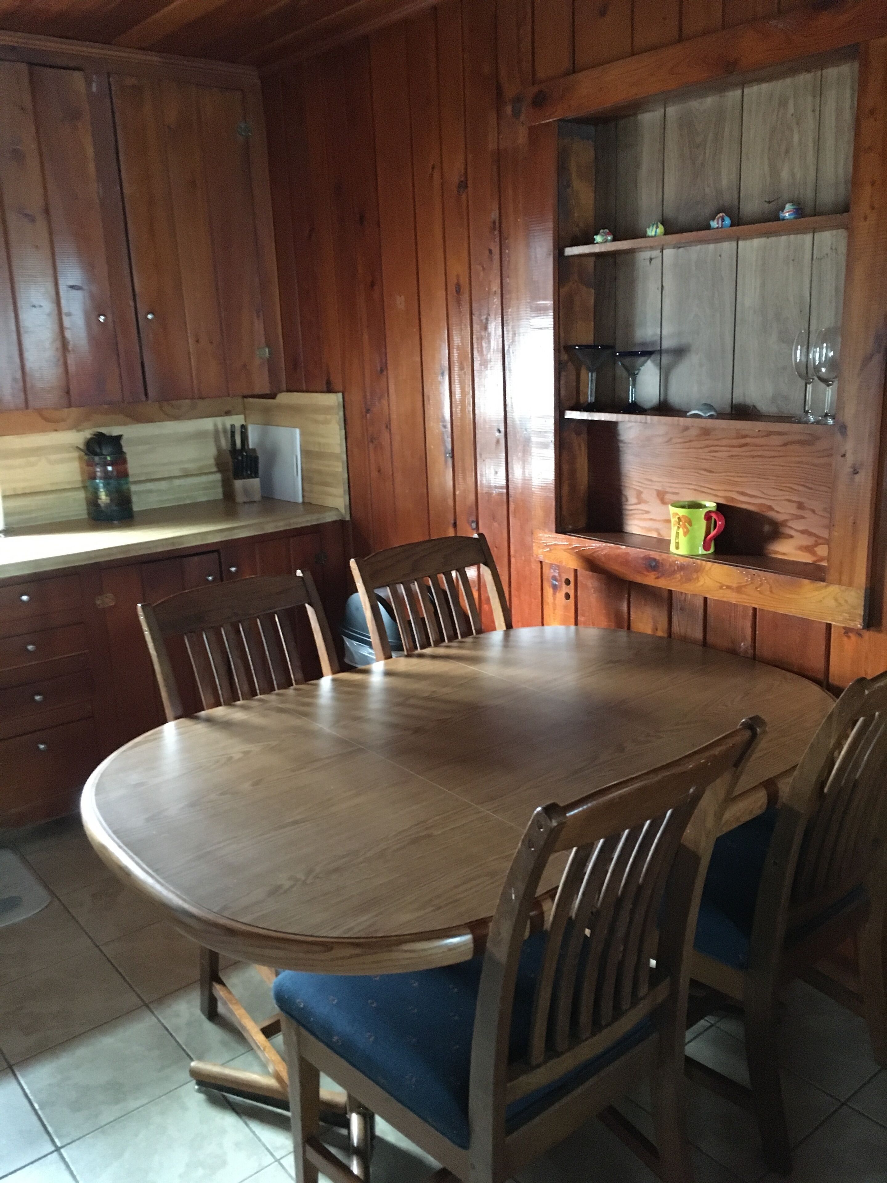 A wooden dining table with chairs, a kitchen area with wooden cabinets, and a shelf with glasses.