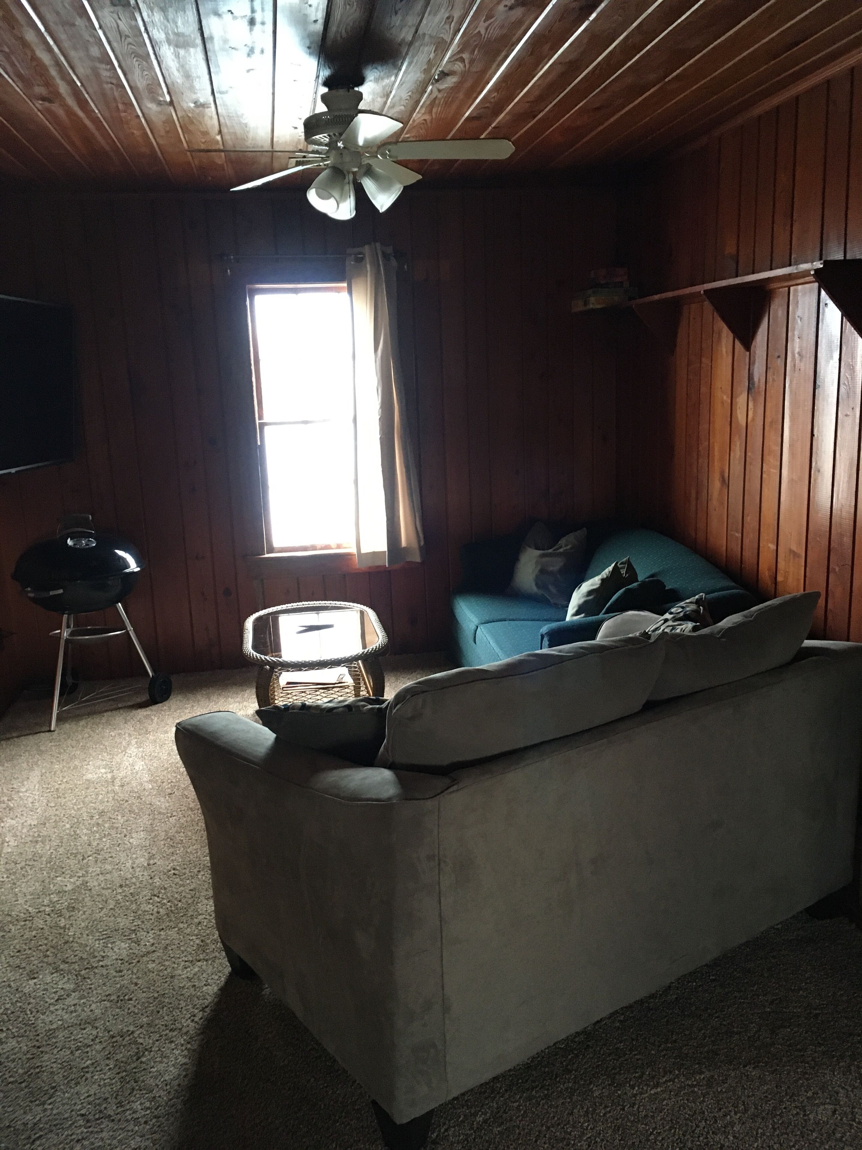 A room with a wooden ceiling, a ceiling fan, a window with curtains, a sofa, a small table, and a television mounted on the wall.