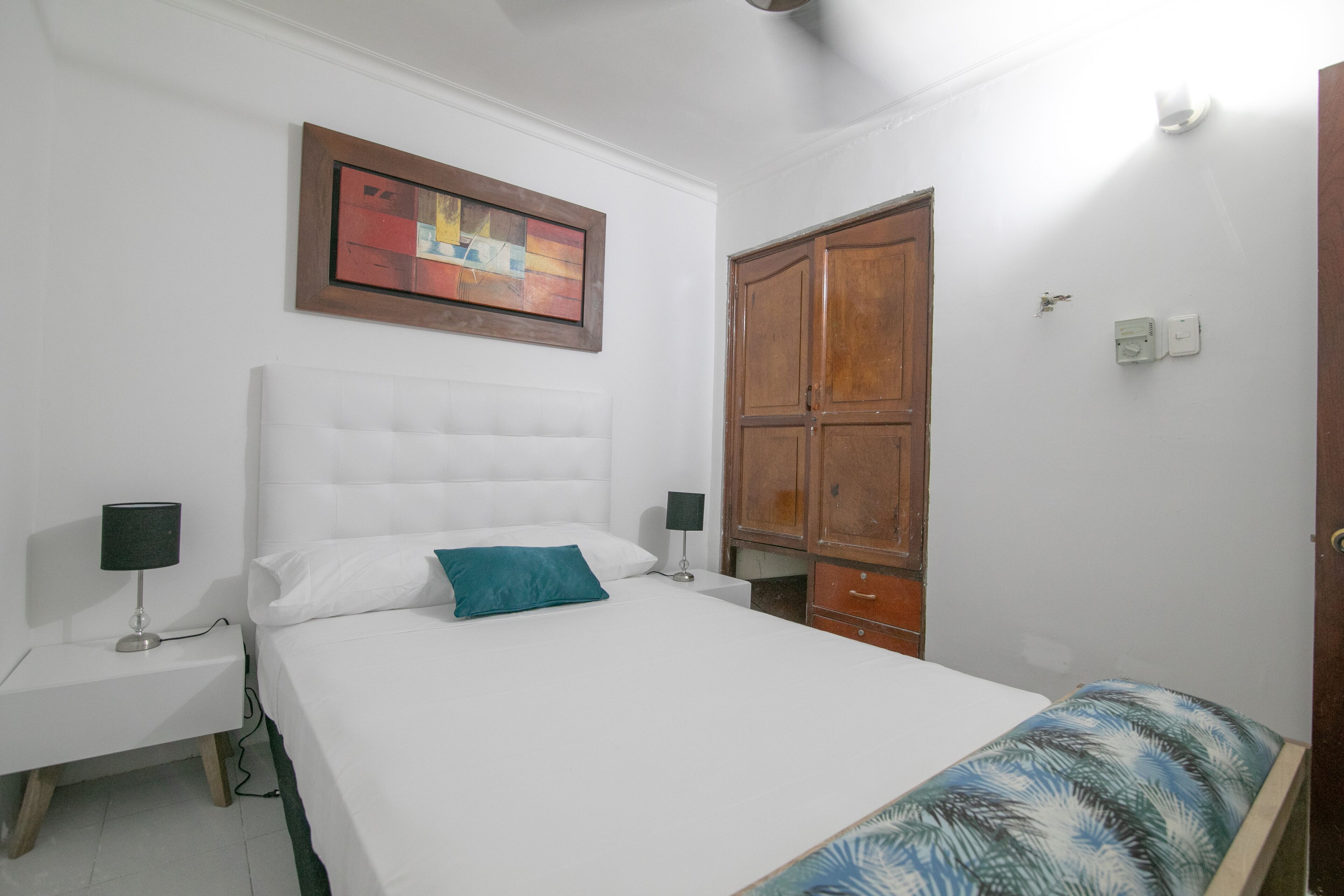 A bedroom with a large bed, two bedside tables with lamps, a wooden wardrobe, and a framed artwork on the wall.
