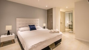 Une chambre moderne avec un grand lit, des tables de chevet, une commode et une salle de bain visible à travers une porte ouverte.