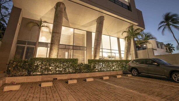 Un bâtiment moderne avec de grandes colonnes, une voiture garée devant et des palmiers en arrière-plan.